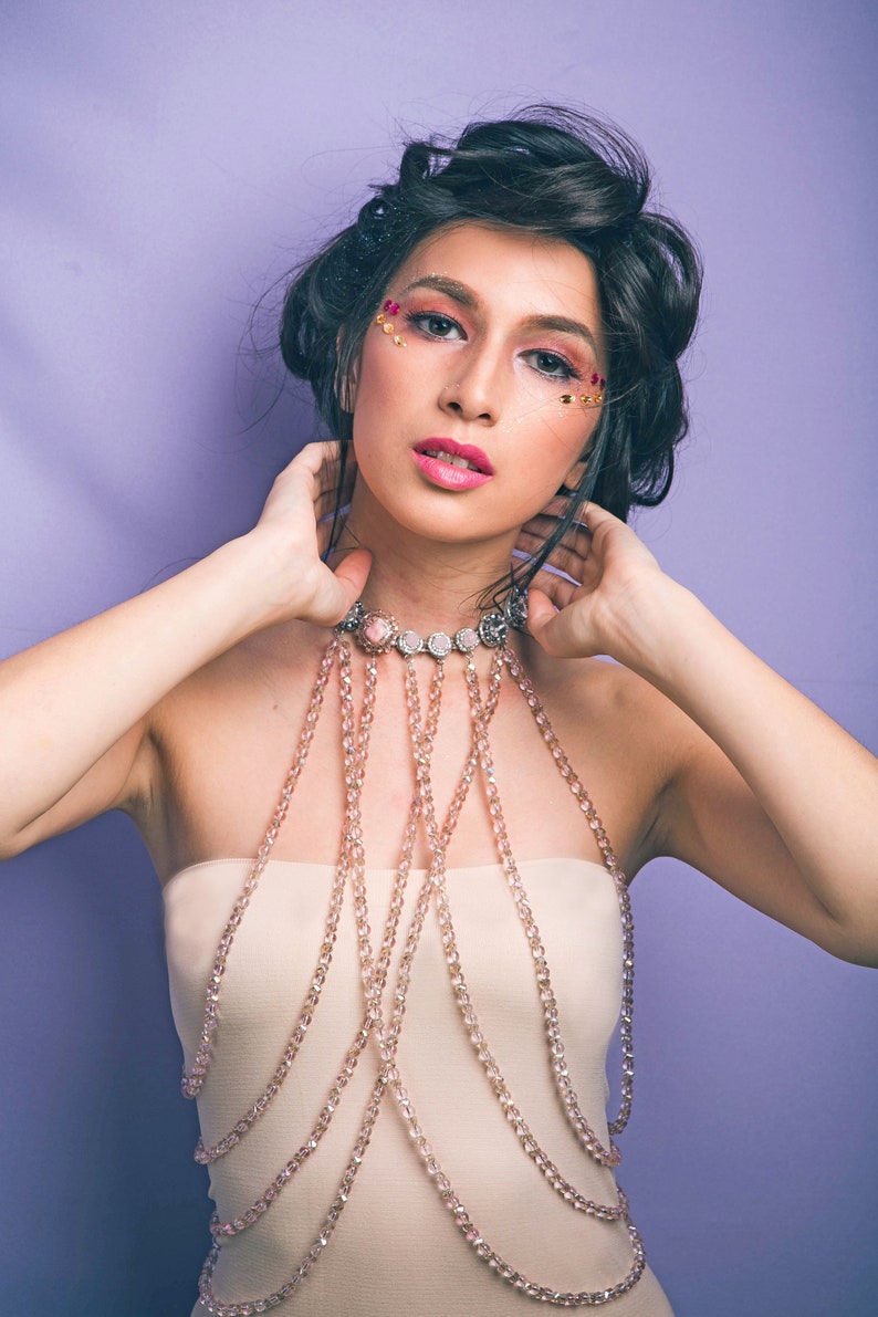 May include: A woman with dark hair and makeup featuring pink and gold glitter is wearing a delicate, multi-layered beaded necklace. The necklace is made of small, clear beads and has a silver clasp. The woman is wearing a strapless top and has her hands on her neck.