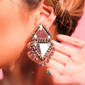 May include: Silver-toned geometric earrings with a mirrored surface and dangling beads. The earrings are a diamond shape with a triangle at the top and bottom. The earrings are being held up to the ear.