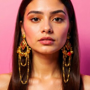 May include: Gold-toned chandelier earrings with red and clear accents. The earrings feature a diamond-shaped top with dangling chains and beads. The background is a solid pink.
