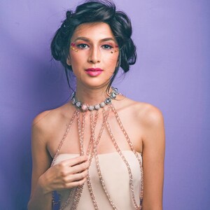 May include: A woman with dark hair and makeup featuring rhinestones is wearing a layered beaded necklace with a large pendant. The necklace is a light pink color and is worn over a strapless top.