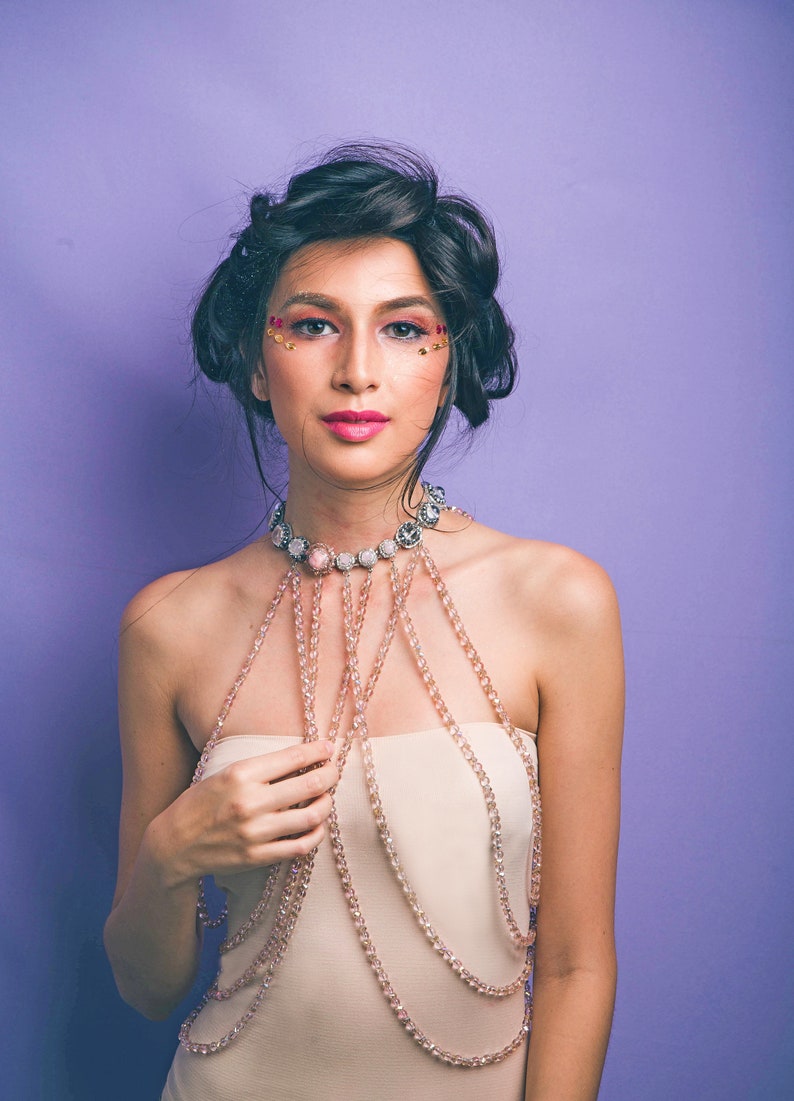 May include: A woman with dark hair and makeup featuring rhinestones is wearing a layered beaded necklace with a large pendant. The necklace is a light pink color and is worn over a strapless top.