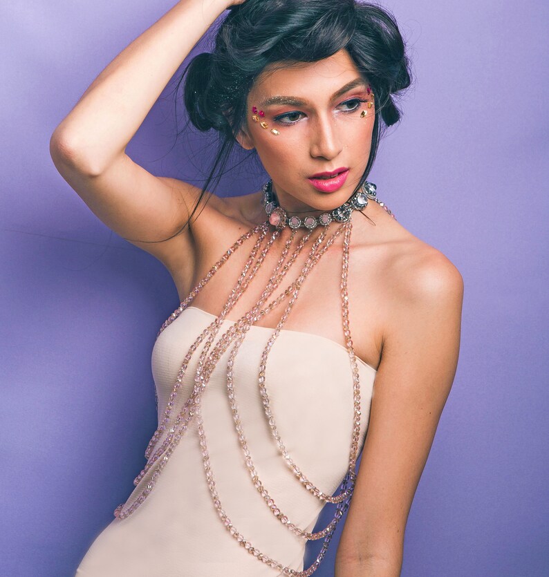 May include: A woman with dark hair and sparkling makeup wears a white strapless top with a layered beaded necklace. The necklace is made of multiple strands of small, clear beads.
