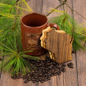 May include: A brown ceramic mug with a design on the side sits on a wooden coaster shaped like the state of Wisconsin. The coaster is made of multiple layers of wood and is surrounded by coffee beans.