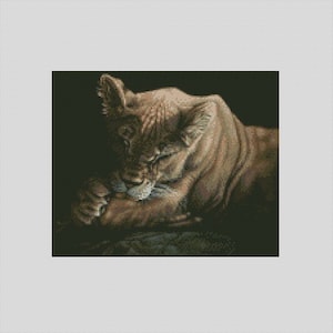 May include: A detailed cross-stitch pattern of a lioness, rendered in shades of brown and tan. The lioness is depicted in a relaxed pose, with her head resting on her paws. The background is a dark, solid color, which highlights the lioness.