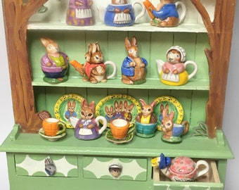 Könnte beinhalten: Ein hellgrüner Holzschrank mit gemalten Baummotiven, ausgestattet mit drei Regalen voller Miniatur-Keramikhasen, Teekannen, Tassen und Tellern. Der Sockel enthält drei Schubladen mit kleinen tierförmigen Knöpfen und einer geblümten Teekanne.