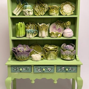 May include: A light green wooden hutch with three drawers featuring painted vine motifs, filled with miniature ceramic vegetable-shaped dishes. The shelves display various sculpted cabbage, lettuce, and cauliflower pieces in shades of green and purple.