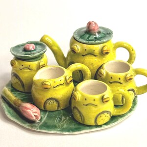 Puede incluir: Un juego de té de cerámica amarillo brillante, que incluye una tetera, una jarra de crema, un azucarero y dos tazas. Cada pieza está adornada con caras de rana y descansa sobre una bandeja verde con forma de hoja. Una flor rosa añade un toque de color.