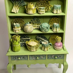 May include: A light green miniature cabinet with three drawers, displaying ceramic vegetables. The cabinet has a scalloped top and curved legs. The shelves hold ceramic leeks, cabbage, artichokes, and other vegetable-shaped items.
