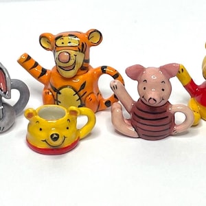 Può includere: Un set da tè a tema Winnie the Pooh con una teiera a forma di asino grigio, una teiera a forma di tigre arancione e gialla, una teiera a forma di maiale rosa, una tazza gialla con la faccia di un orso e una teiera gialla con la faccia di un orso.