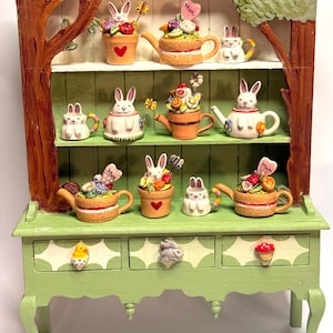 May include: A green cabinet with three drawers and shelves, decorated with bunny and floral figurines. The cabinet has a tree design on the sides and is painted in green and brown. The figurines include teapots and flower pots, perfect for Easter.