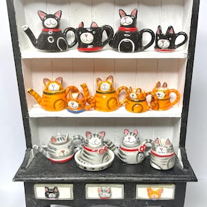 May include: A black wooden hutch with three shelves displays a collection of ceramic cat teapots. The teapots are in various colours, including black, orange, and grey. The hutch has two drawers with cat faces on the fronts. The bottom shelf has a few more cat teapots.