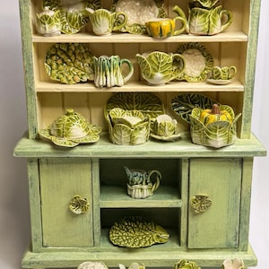 May include: A miniature green cabinet with shelves displaying a collection of ceramic dishware. The dishware includes teacups, teapots, plates, and serving dishes, all designed to resemble cabbage leaves and other vegetables. The cabinet is painted in a light green color.