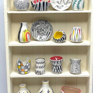 Può includere: Una collezione di piccoli oggetti in ceramica dipinti a mano esposti su una mensola bianca. I pezzi includono vasi, ciotole e piatti con vari motivi, tra cui pois e strisce in bianco e nero e disegni colorati.