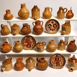 May include: A collection of miniature brown ceramic jugs, bottles, and decorative plates arranged on a white tiered shelf. The items feature moulded bearded faces, cork stoppers, and hand-painted illustrations including a mermaid and historical figures.