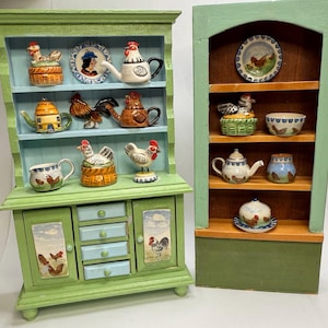 Puede incluir: Dos armarios verdes en miniatura con estantes, adornados con figuritas de gallinas de cerámica, teteras y platos. Los armarios tienen una estética rústica de granja, con detalles pintados y un encantador diseño de estilo campestre. Los armarios están pintados en tonos verdes y azules.