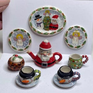 May include: A miniature Christmas-themed tea set. The set includes a Santa teapot, snowman cups, and plates with angel and snowman designs. The set is painted with bright colors and festive details, perfect for dollhouses or holiday decor.