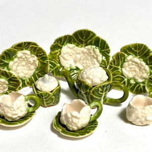 May include: A miniature ceramic tea set with a cauliflower theme. The set includes a teapot, cups, saucers, a sugar bowl, and plates, all in white and green. The pieces are shaped like cauliflower and cabbage leaves.