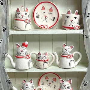 Może przedstawiać: Biały ceramiczny zestaw do herbaty z czajnikami, filiżankami i spodkami w kształcie kota. Zestaw jest ozdobiony czerwonymi i czarnymi detalami, w tym czerwoną muszką i czarnymi wąsami na każdym kocie.