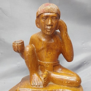 May include: A vintage wooden sculpture of a seated figure, holding a small cup and resting an arm on its knee. The figure is carved from wood and sits atop a round, tiered base. The sculpture has a warm, brown color.