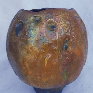 May include: A handcrafted, iridescent ceramic vase with a textured surface. The vase has a bulbous shape with a dark, textured base and rim. The exterior features a blend of bronze, gold, and blue hues, with circular indentations adding visual interest.