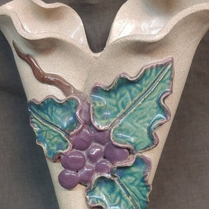 May include: A beige ceramic wall vase with a V-shape design. It features a cluster of purple grapes and green leaves with teal edges, along with brown stems. The vase has a textured surface.
