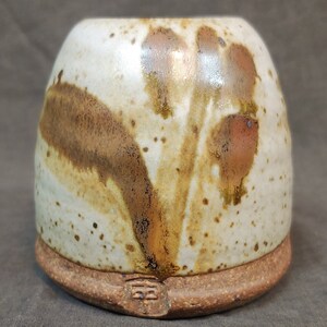 May include: A handmade ceramic mug with a speckled off-white glaze and brown and rust-colored abstract designs. The base is a darker, unglazed terracotta color. The mug has a unique, organic shape and a small, stamped mark on the base.