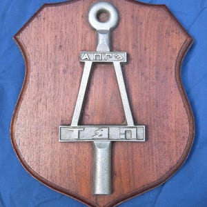May include: A shield-shaped wooden plaque featuring a central silver-toned metal emblem. The emblem consists of a vertical rod with a circular top, a triangular frame, and a horizontal bar displaying the embossed Cyrillic characters АПГЭ and ТЯП.