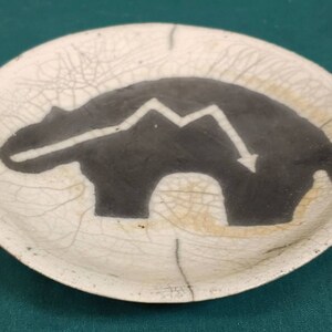 May include: A small, round, off-white ceramic dish with a black bear silhouette and a white downward arrow design. The dish has a cracked, aged appearance.