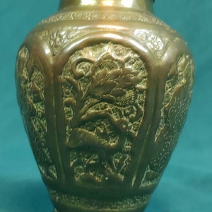 May include: Bronze-colored vase with detailed relief carvings. The vase features floral and animal motifs, with a slightly flared base and a narrow neck. The surface has a patina, suggesting age. The overall design is ornate and decorative.