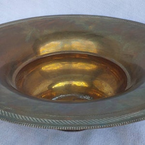 May include: A large, antique brass bowl with a wide, flat rim and a deep, rounded center. The rim features a decorative, textured border. The bowl has a warm, golden hue and shows signs of age.