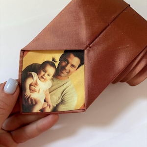 Pode incluir: Uma gravata de seda marrom com um inserto de foto quadrado que apresenta um homem e uma criança pequena. A foto é de um homem segurando uma menina pequena nos braços. A foto é impressa em um fundo amarelo claro.