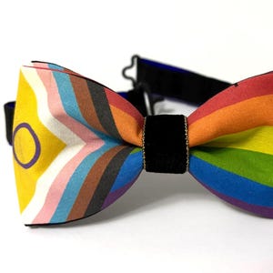 May include: A rainbow striped bow tie with a black velvet band and gold trim. The bow tie has a yellow circle with a purple ring in the center.