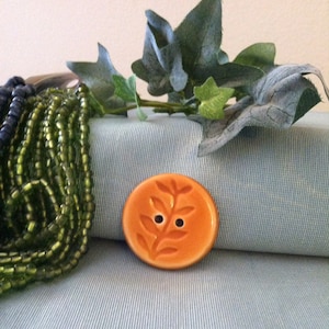 May include: A round, orange ceramic button with a raised design of a leafy branch and two small holes. The button is sitting on a light blue fabric surface.