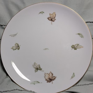 May include: White ceramic plate with a gold rim, decorated with scattered autumn leaves in shades of brown, green, and beige. The plate has a simple, elegant design, suitable for serving food.