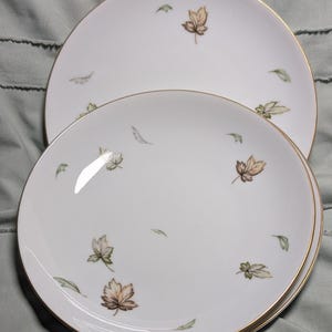 May include: White ceramic plates with a gold rim and a scattered leaf design in shades of brown and green. The plates are of different sizes and are stacked. The plates are suitable for serving food.