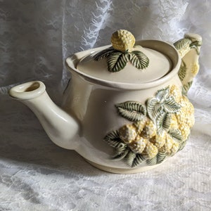 May include: A white ceramic teapot with a lid and a handle. The teapot is decorated with a raised design of green leaves and yellow berries.