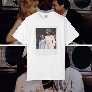 Può includere: Una t-shirt bianca a maniche corte con una stampa grafica rettangolare centrata. La stampa mostra una scena fotografica di un bambino con un cappotto azzurro e una donna con un cappotto bianco, incorniciata da un bordo bianco stile polaroid.