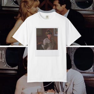 Può includere: Una t-shirt bianca a maniche corte con una stampa grafica rettangolare centrata. La stampa mostra una fotografia in stile vintage di una persona che indossa occhiali da sole scuri e una camicia a colletto chiaro, tenendo una sigaretta.