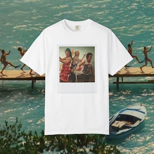 Könnte beinhalten: Ein weißes Kurzarm-T-Shirt mit einem mittigen, rechteckigen Grafikdruck. Der Druck zeigt drei Personen in einem Innenraum, wobei eine Person ein Handtuch auf dem Kopf und ein rotes gemustertes Kleidungsstück trägt, eine andere ein weißes T-Shirt.