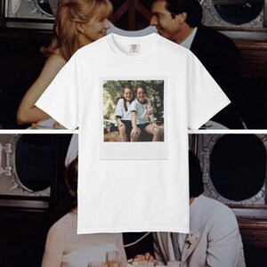 Può includere: Una t-shirt bianca a maniche corte con una stampa grafica rettangolare centrata che raffigura una fotografia in stile Polaroid vintage. L'immagine mostra due persone con lunghi capelli castani che indossano camicie bianche e pantaloncini scuri.