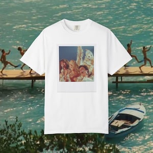 Op de afbeelding: Een wit t-shirt met korte mouwen en een gecentreerde rechthoekige grafische print van drie personen die op een podium in microfoons zingen. De print is vormgegeven als een vintage polaroidfoto met een witte rand op het t-shirt.