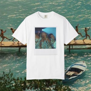 以下が含まれることがあります： 中央に長方形のグラフィックプリントが施された白い半袖Tシャツ。ステージの青い照明の下でマイクに向かって歌う、きらびやかな衣装を着た3人の人物が写っており、ヴィンテージのポラロイド写真のようなスタイルでデザインされています。