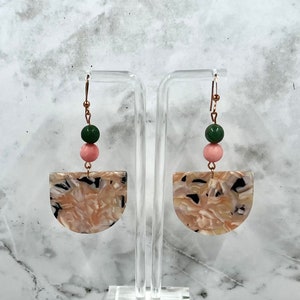 May include: A pair of gold hoop earrings with a pink and green bead dangle. The dangle features a half-circle shaped resin piece with a marbled pattern.