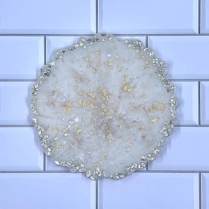 May include: A round, white resin coaster with gold leaf accents and a silver glitter edge. The coaster is set against a white tile background, showcasing its decorative design. This handmade item is perfect for home decor.