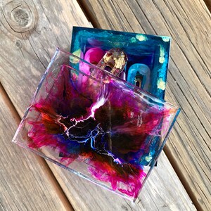 May include: A clear resin box with a vibrant abstract design in shades of pink, purple, blue, and orange. The box contains three decorative items: a pink, a gold, and a blue object. The box sits on a wooden surface.