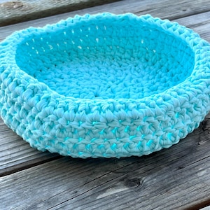 May include: A round, light blue crochet basket. The basket is made of thick, textured yarn and sits on a weathered wooden surface. The basket's sides are approximately 6 inches in diameter and 3 inches tall.