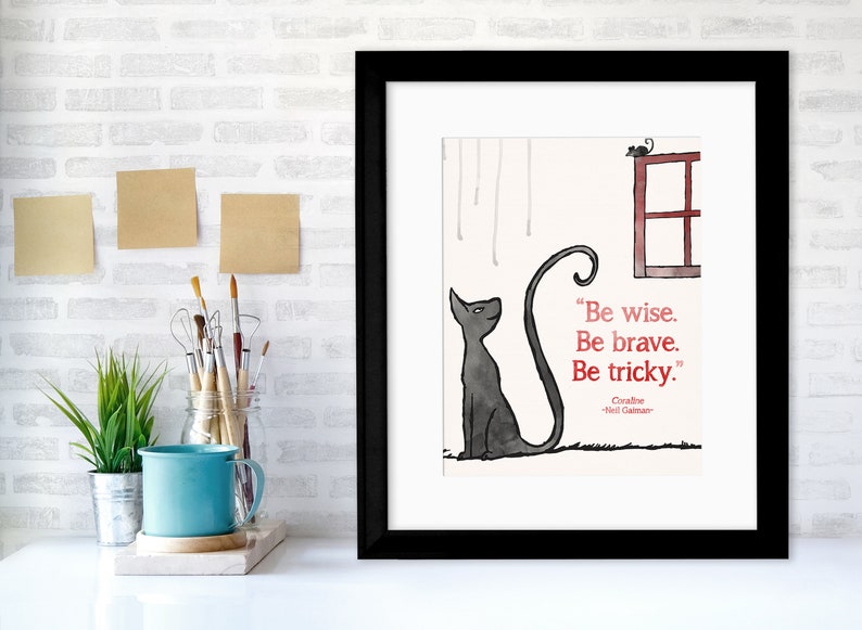 Puede incluir: Una impresi&oacute;n enmarcada con un gato negro sentado y mirando hacia arriba a una ventana roja con un peque&ntilde;o rat&oacute;n en la parte superior. La cita "S&eacute; sabio. S&eacute; valiente. S&eacute; astuto." est&aacute; escrita en rojo debajo del gato. La cita se atribuye a "Coraline" y "Neil Gaiman".