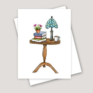 May include: A wooden end table with a curved leg design. On the table is a stack of books, a small potted plant with pink flowers, a blue and green lamp with a black base, and a white mug.