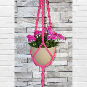 Könnte beinhalten: Ein rosa Makramee-Pflanzenhänger mit einem beigefarbenen Topf und rosa Blumen, die an einer weißen Ziegel- und Steinwand hängen.