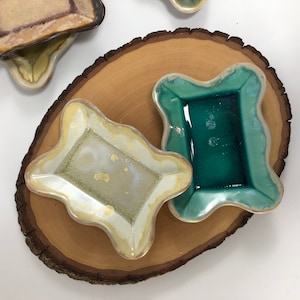 May include: Three ceramic dishes with irregular edges, one is green, one is white with gold accents, and one is a light brown colour. The dishes are on a wooden slice.
